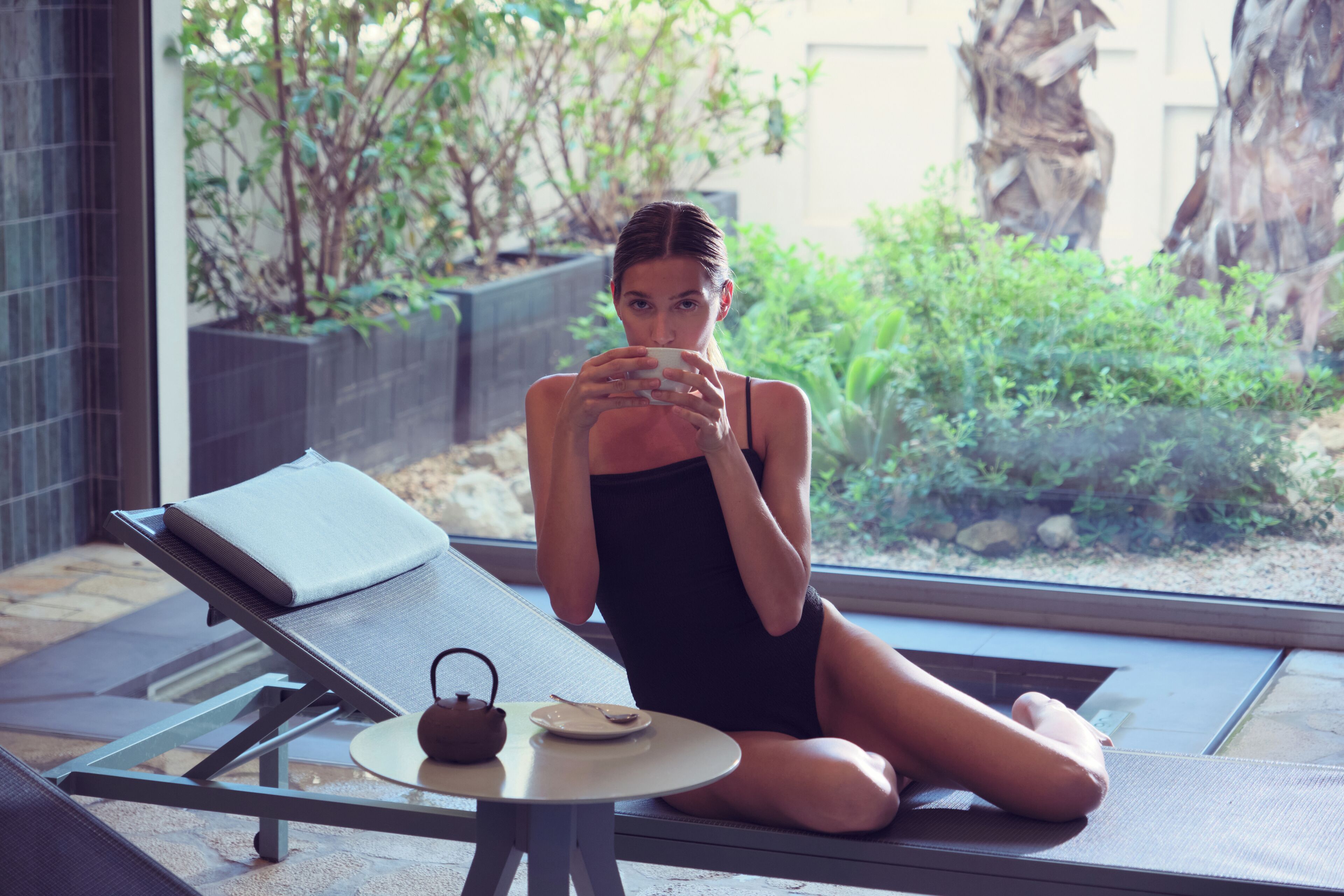 A person sitting on a sun lounger with a table beside them, a small teapot, and a plate.
