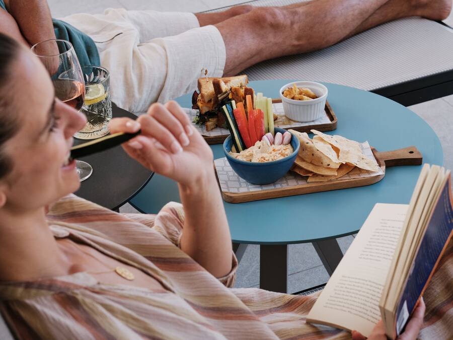 Iemand leest een boek aan een ronde tafel met een maaltijd bestaande uit een belegd broodje, groenten en chips.