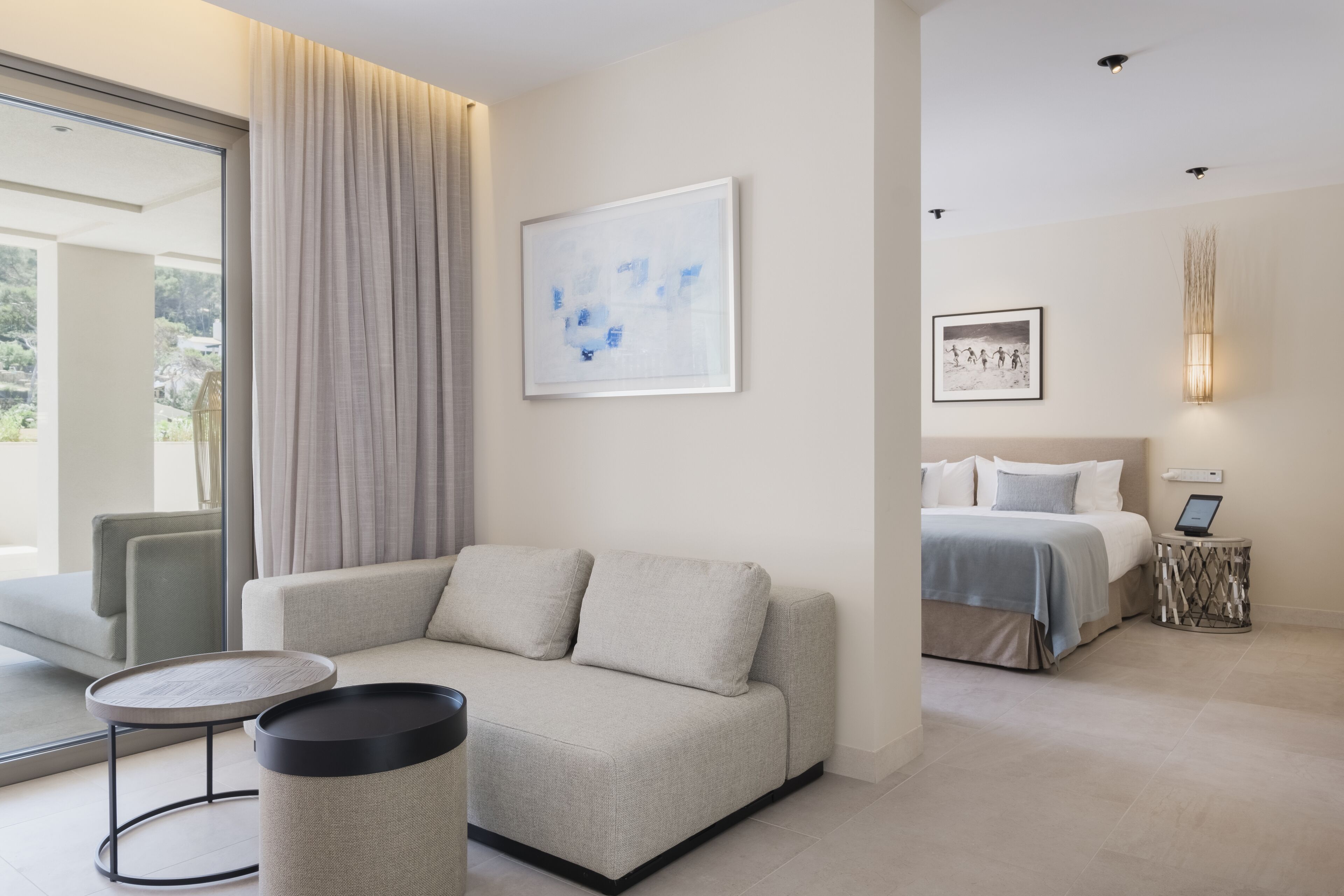 A modern hotel room with a sofa, a round table, a bed with a bedside table, and artwork on the walls.