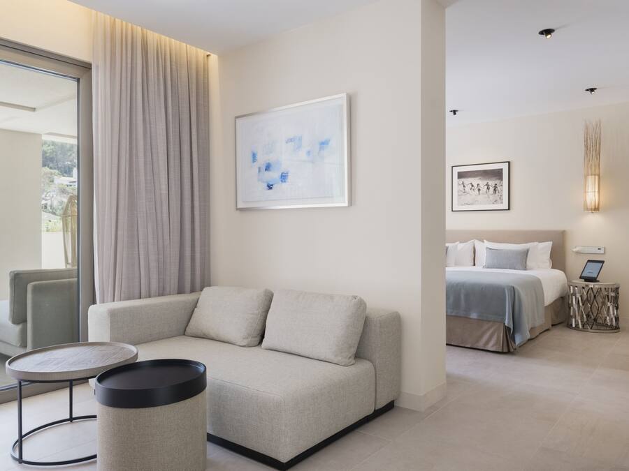 A modern hotel room with a sofa, a round table, a bed with a bedside table, and artwork on the walls.
