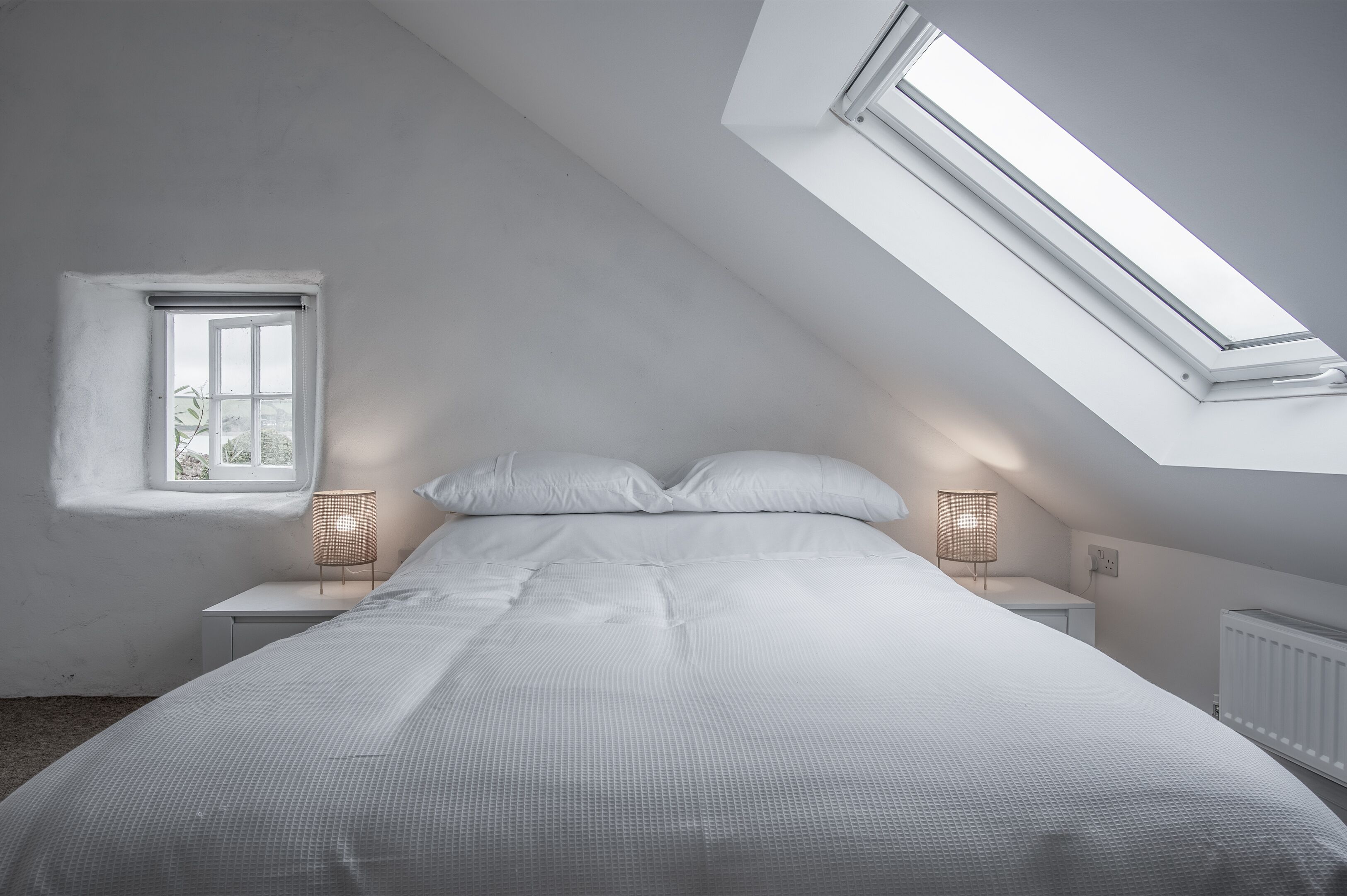 A bedroom with a sloped ceiling, a skylight, a bed, bedside lamps, and a window.