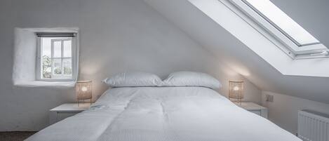 A bedroom with a sloped ceiling, a skylight, a bed, bedside lamps, and a window.