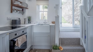 Une cuisine compacte avec des meubles blancs, un four encastré, un évier et une fenêtre donnant sur un mur en pierre.