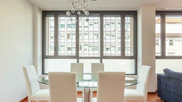 A modern living room with a glass dining table surrounded by white chairs, a blue sofa, and large windows with a city view. - easyhomes - Madonnina view (Milan)