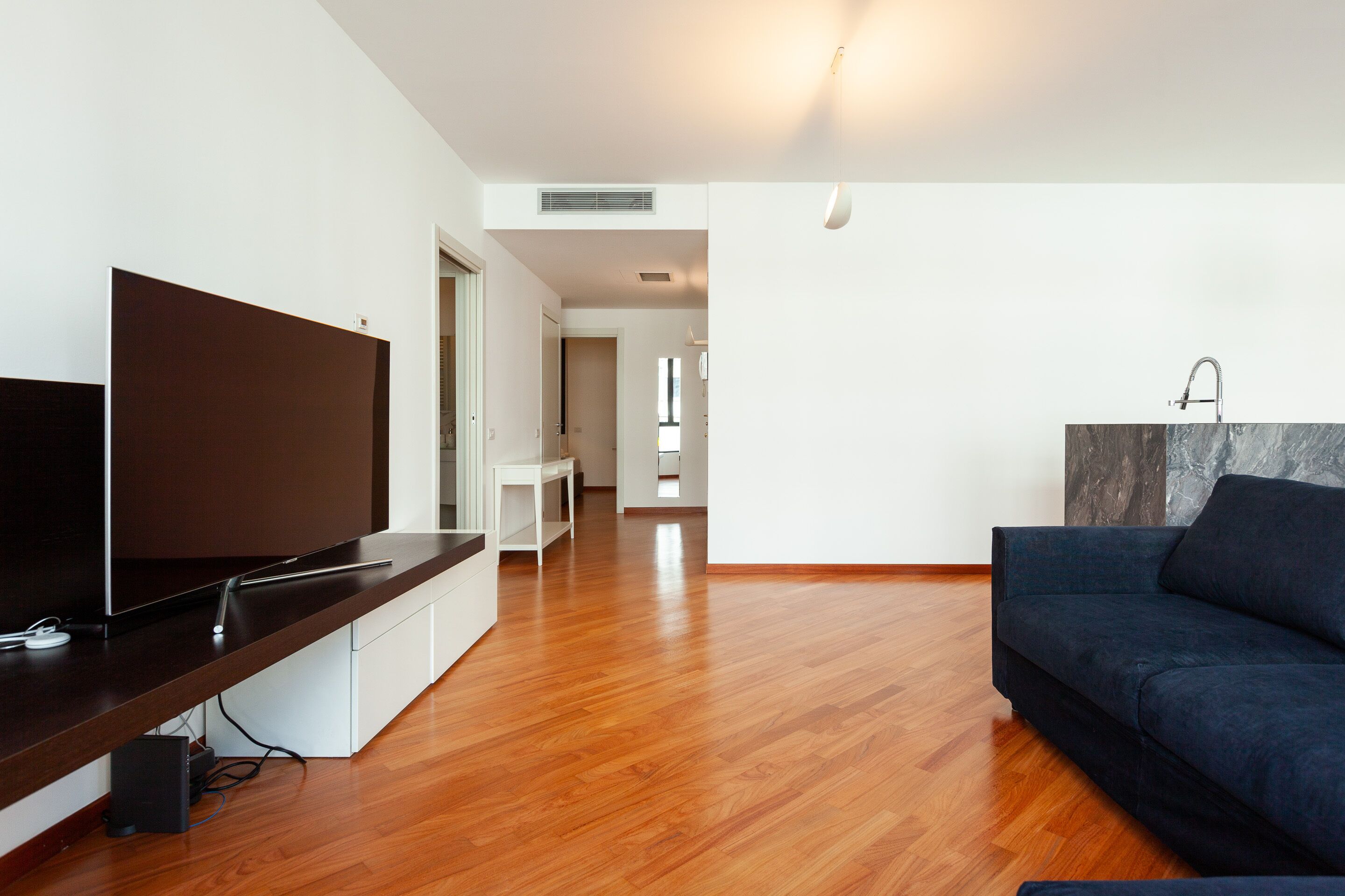 A modern living room with a flat-screen TV, wooden flooring, a dark blue sofa, and a marble accent wall.