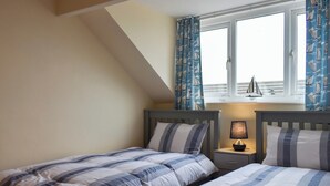 Une chambre avec deux lits, une fenêtre avec des rideaux, une table de chevet avec une lampe et une petite décoration représentant un voilier.