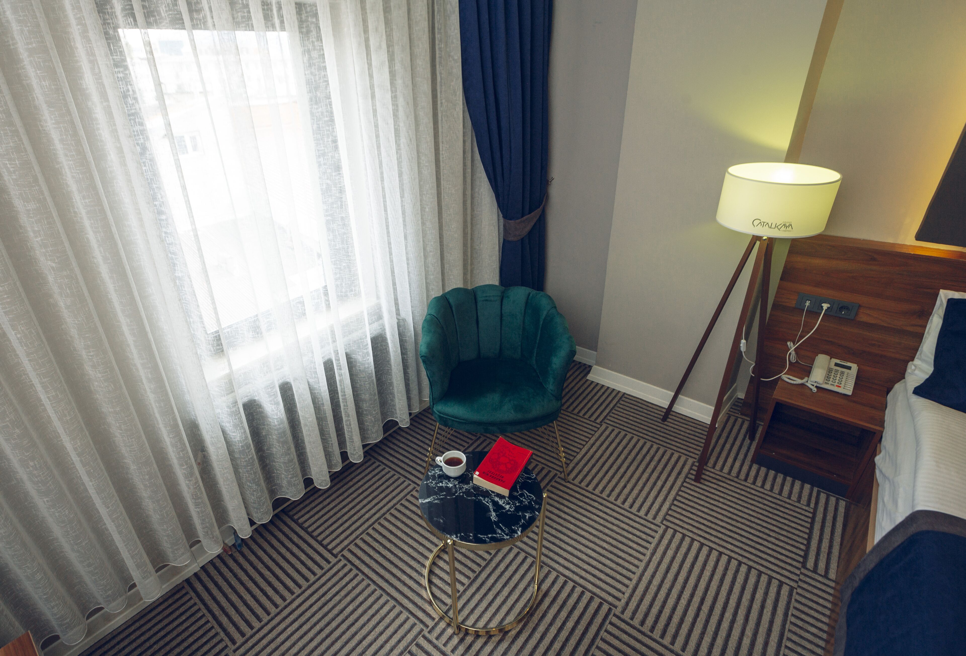 A hotel room with a green chair, a small round table with a book and a cup, a lamp, and a bed.