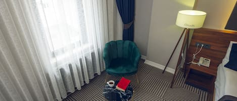 Habitación de hotel con una silla verde, una mesita redonda con un libro y una taza, una lámpara y una cama.