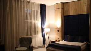 Une chambre d’hôtel avec un lit, une chaise, une petite table, une lampe et une fenêtre avec des rideaux légers.
