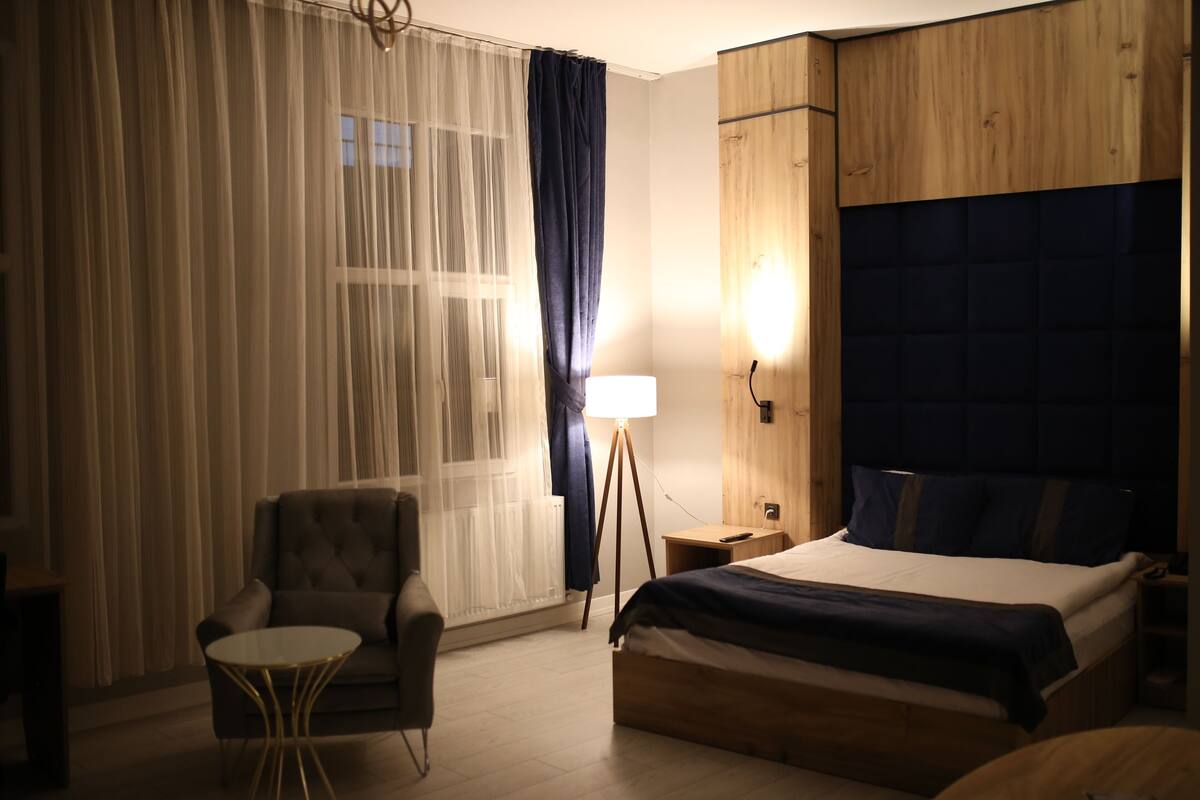 Une chambre d’hôtel avec un lit, une chaise, une petite table, une lampe et une fenêtre avec des rideaux légers.