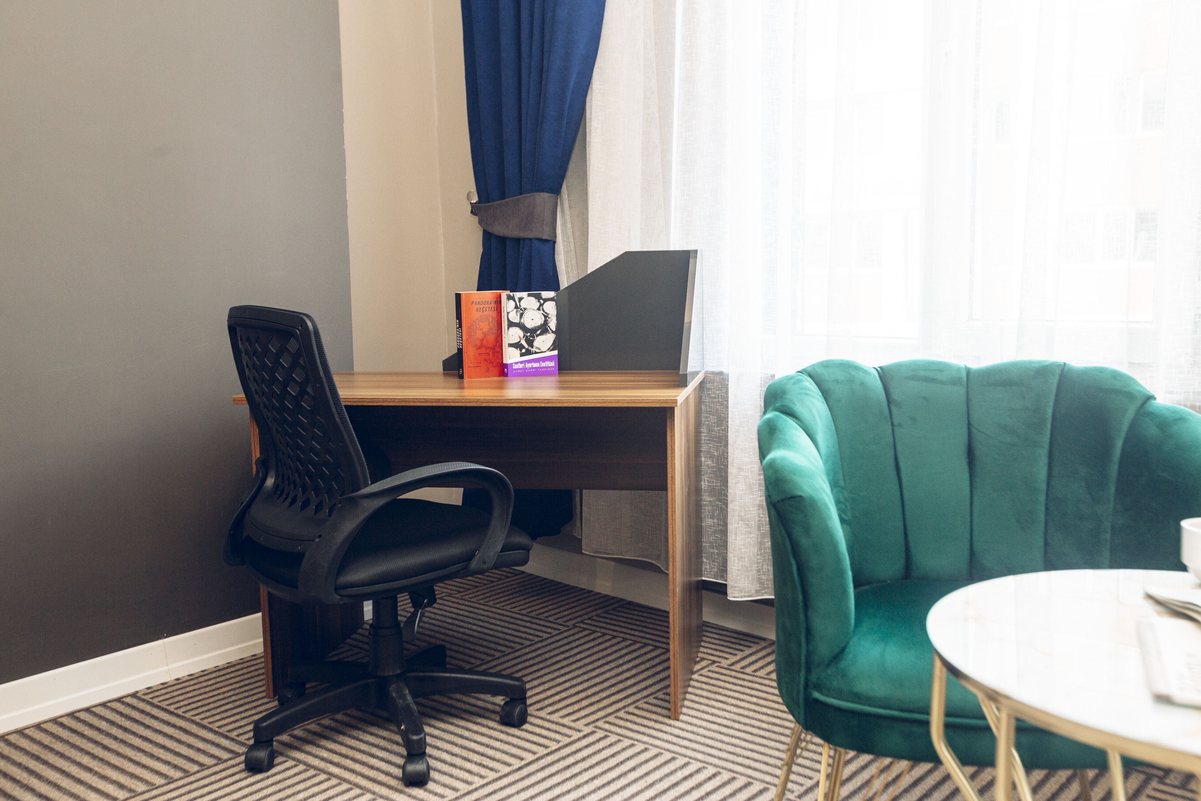 Una habitación con un escritorio, una silla de oficina negra, un sillón azul turquesa y una pequeña mesa redonda.