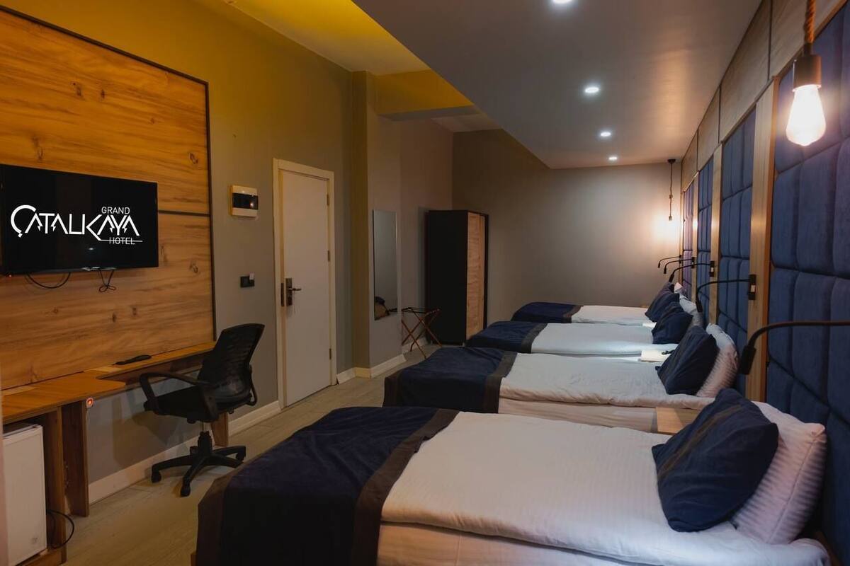 Une chambre d’hôtel avec quatre lits disposés en rangée, un bureau avec une chaise, un téléviseur fixé sur un panneau en bois et une porte.