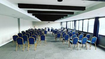 Un salón de conferencias con filas de sillas azules y mesas, ventanas amplias a lo largo de una pared y una alfombra estampada.