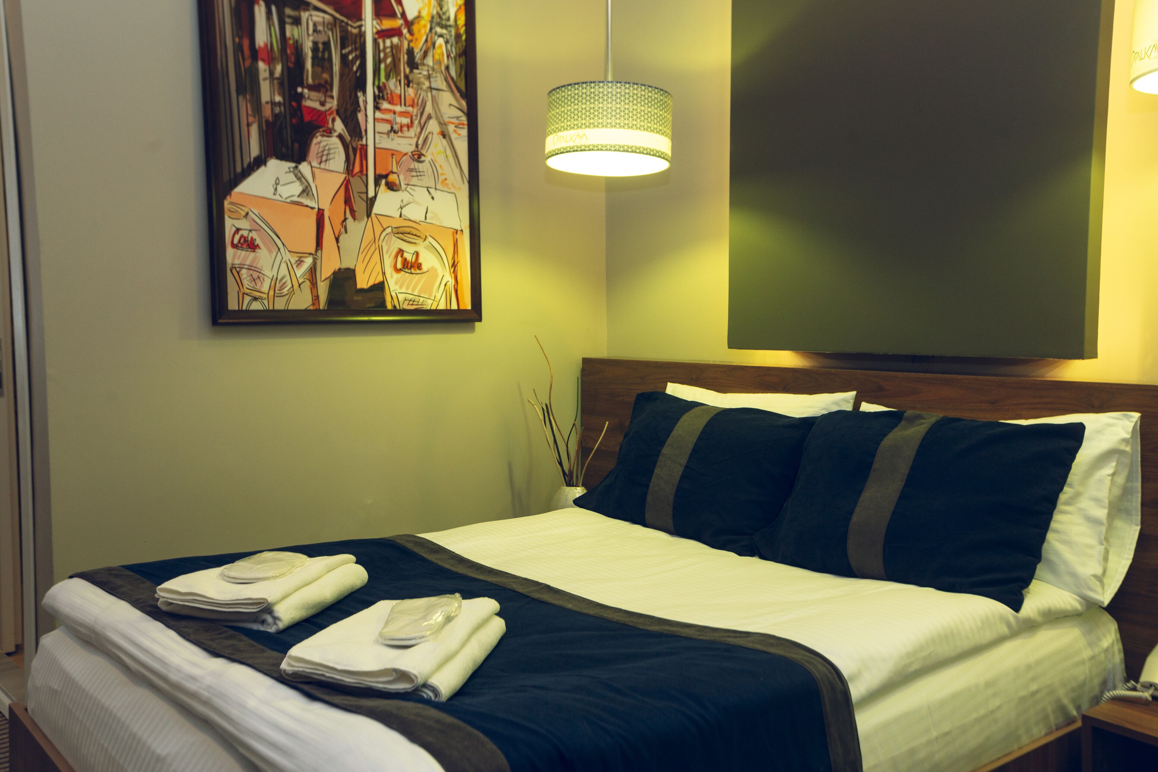 A neatly made bed with a dark blue headboard, white linens, and a blue accent pillow. A framed abstract painting hangs on the wall above the bed.