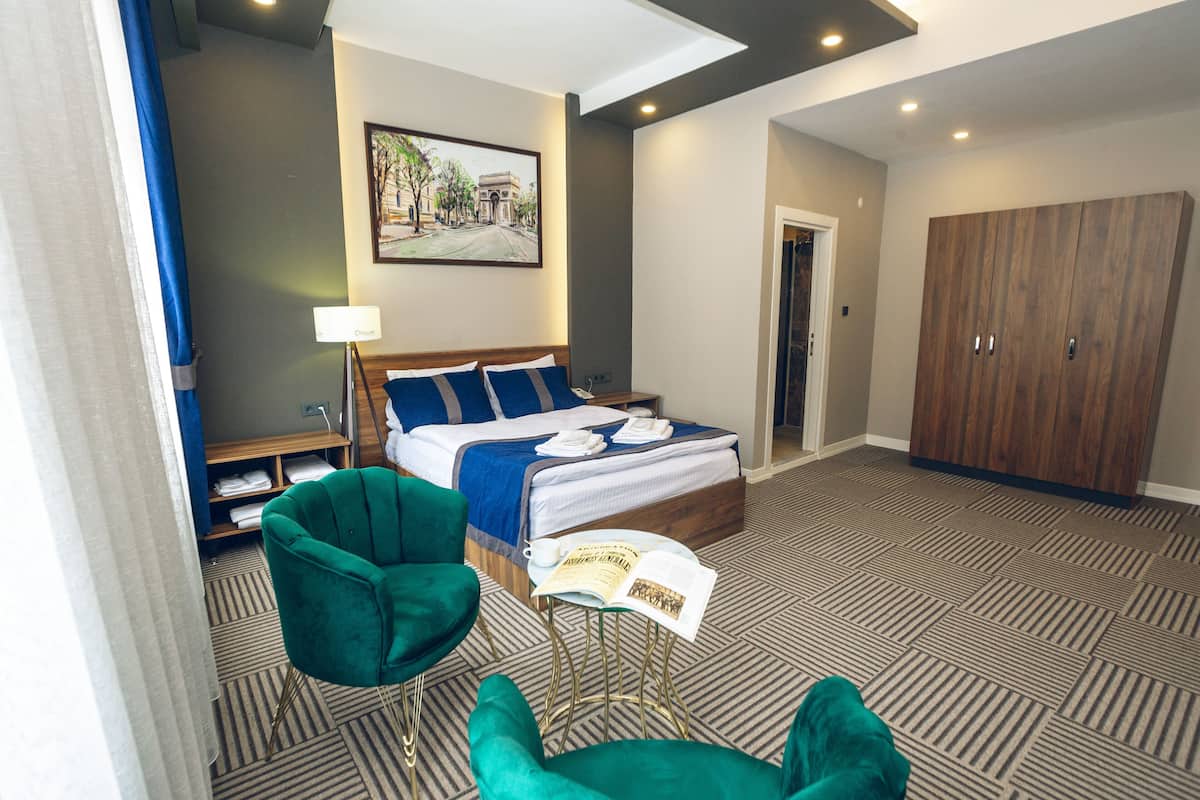 Une chambre d’hôtel avec un lit, deux fauteuils verts, une petite table avec un livre, un tableau au mur et une armoire.