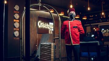 A person in a red uniform standing in a lobby with a luggage rack and clocks displaying times for Istanbul, London, Paris, and Tokyo.