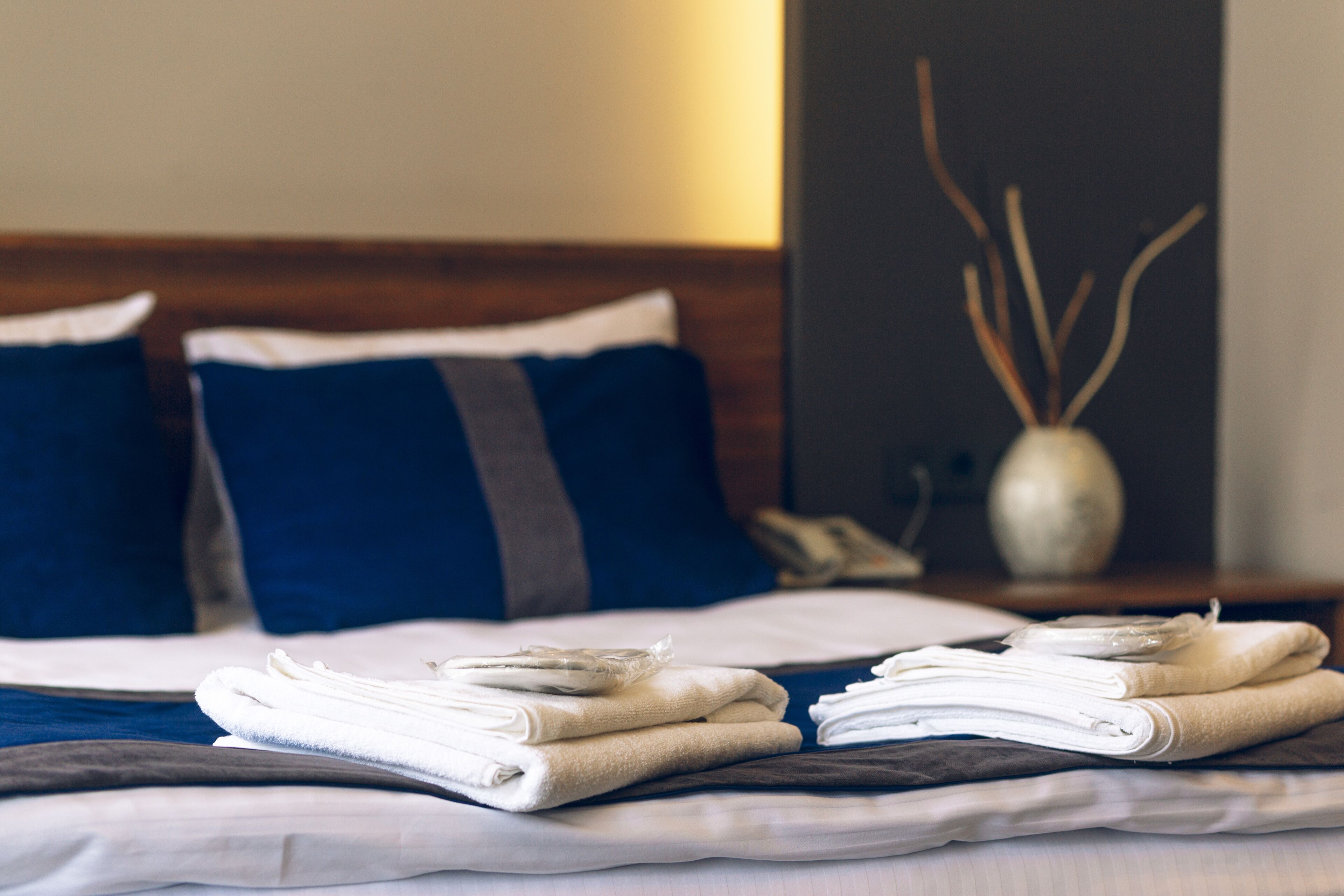 Una cama bien hecha con toallas dobladas y un florero con ramas decorativas sobre una mesita de noche.