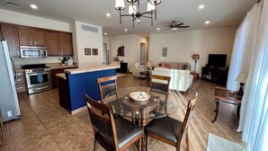 Cozinha moderna com ilha central, área de jantar com mesa de vidro e sala de estar com sofá e televisão.