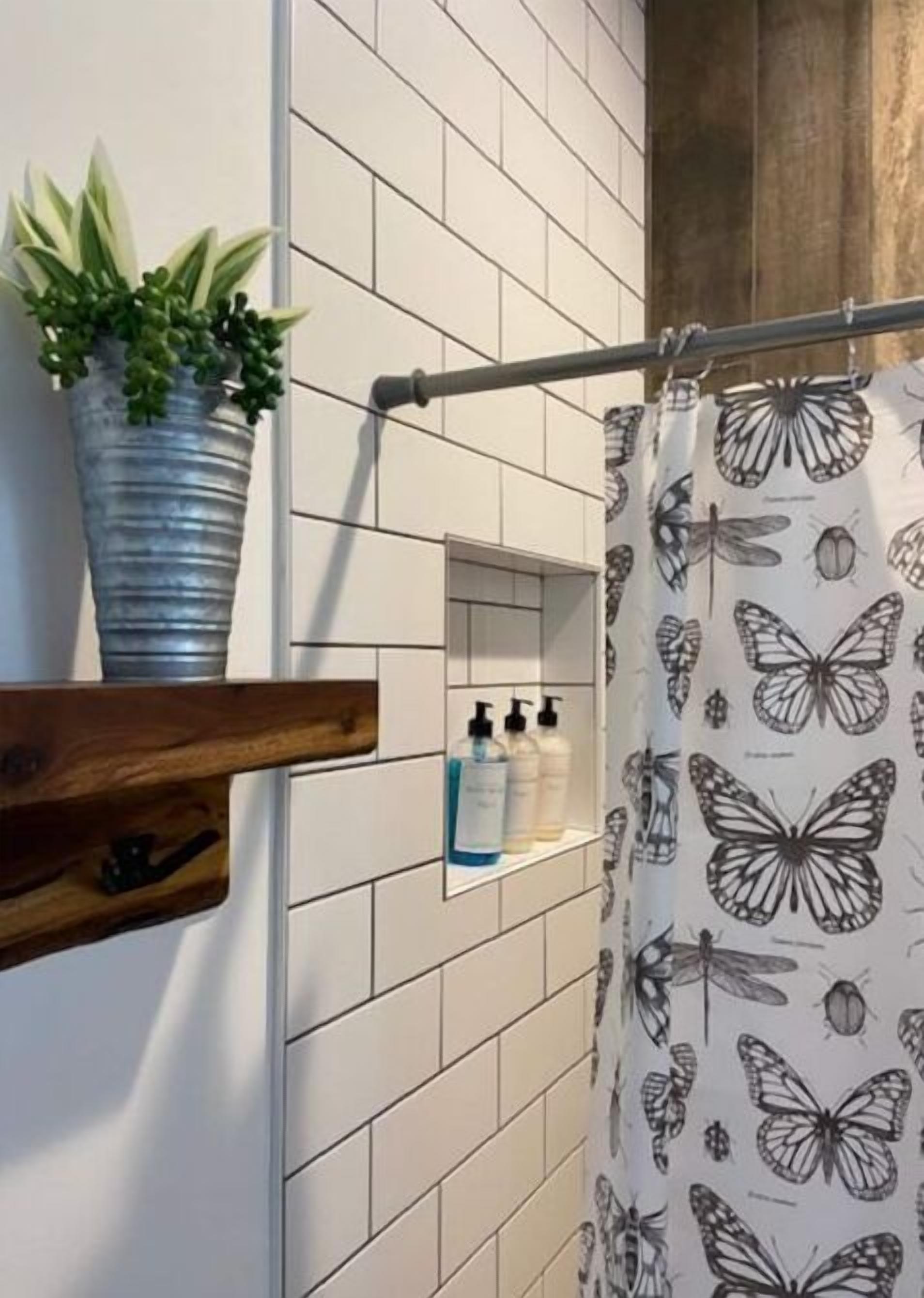 A bathroom with white tiled walls, a shower curtain with butterfly patterns, a potted plant, and a shelf with toiletries.