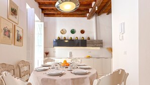 Un comedor con una mesa redonda dispuesta para cuatro personas, sillas blancas, techo de madera y una cocina al fondo.