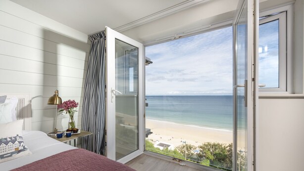 Een slaapkamer met een groot bed, een nachtkastje met een lamp, een vaas met bloemen en uitzicht op het strand door een open deur.