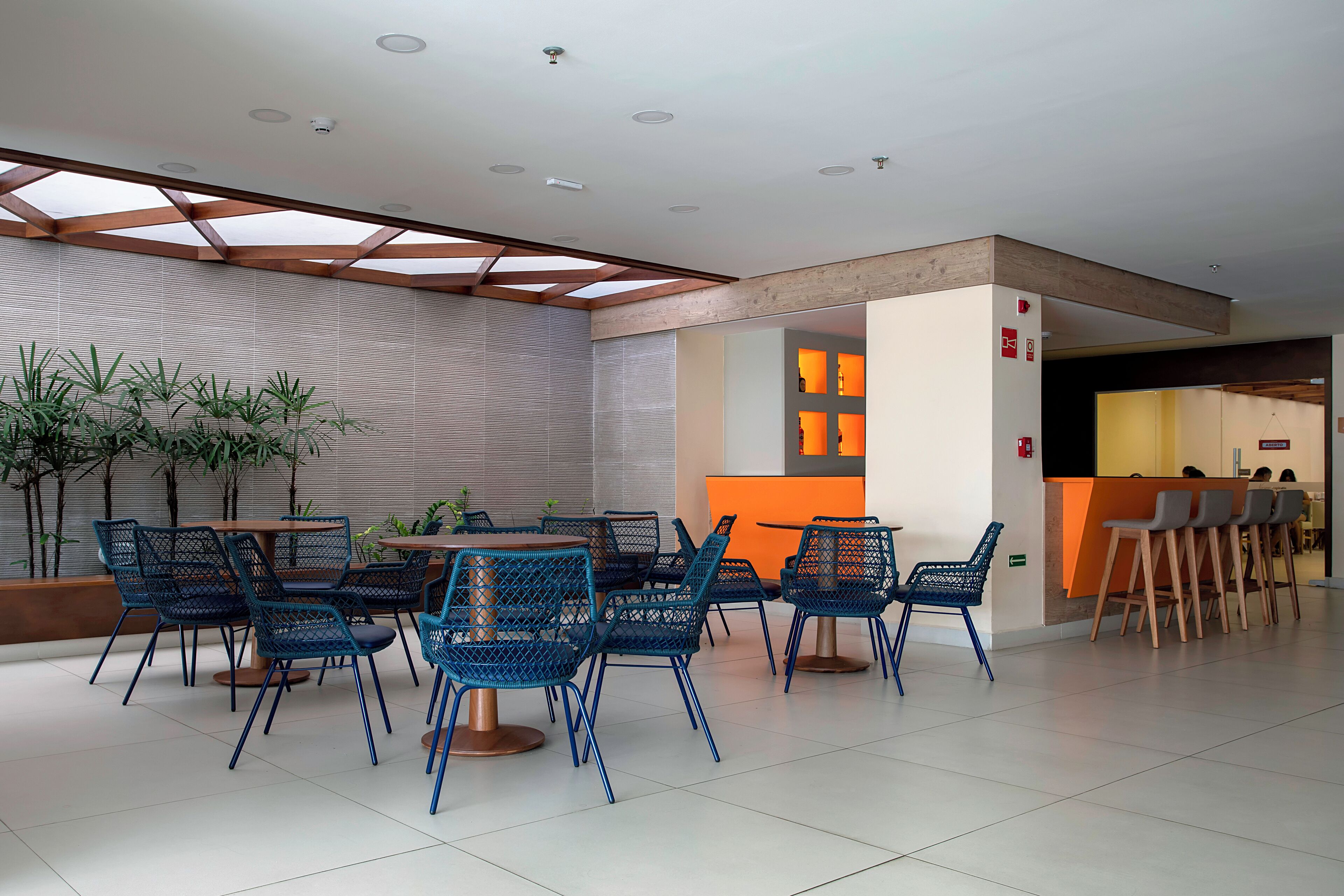 Un espace de restauration intérieur moderne, avec des chaises en treillis métallique bleu et une table ronde, agrémenté d’une verrière et d’un mur aux lignes horizontales.
