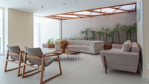 A modern living room with a sectional sofa, two rocking chairs, and a coffee table. There are several potted plants and a large window providing natural light. - Verano Pajuçara by Tropicalis (Maceió)