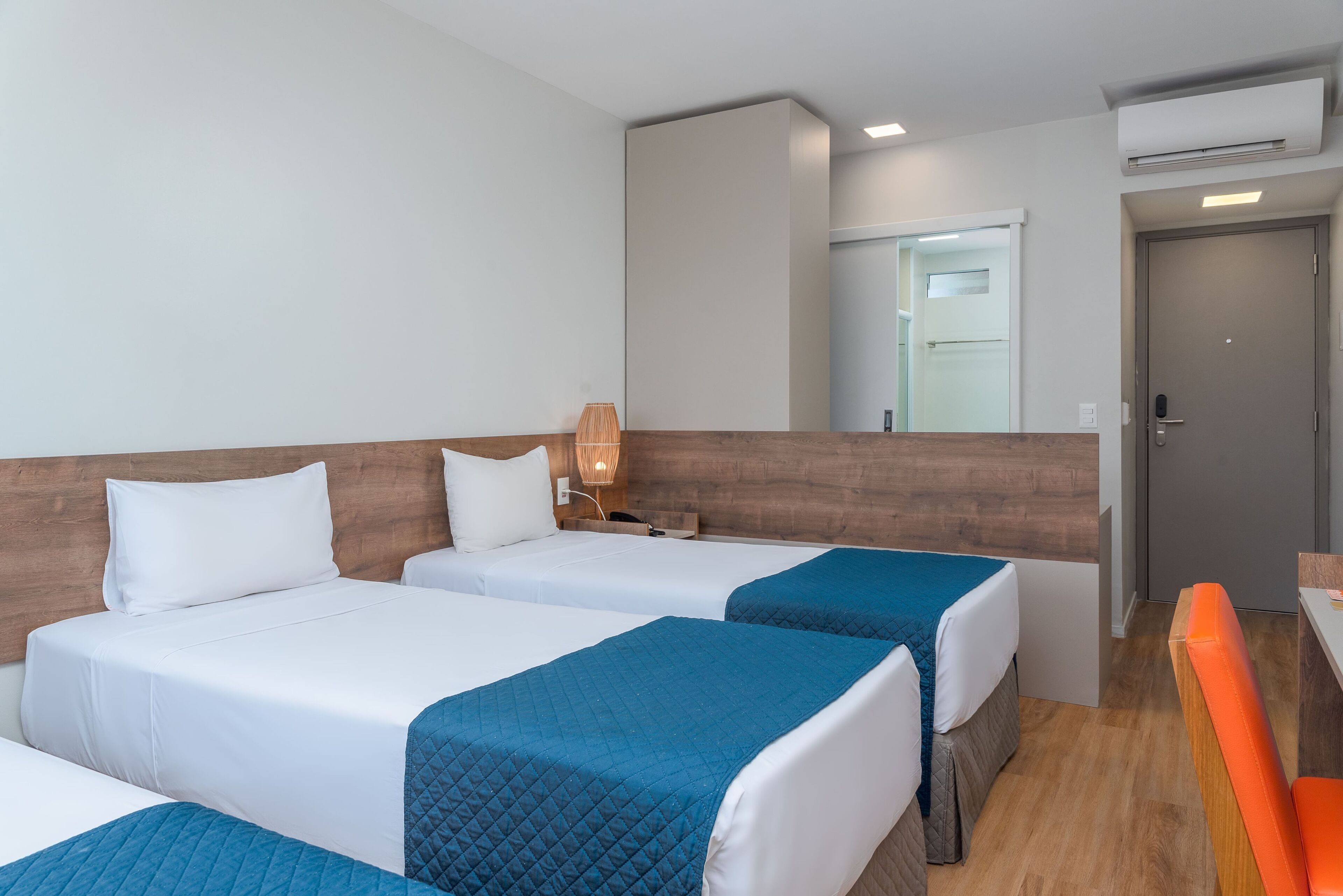 A hotel room with two beds, a wooden headboard, a bedside table with a lamp, and a small seating area with an orange chair.