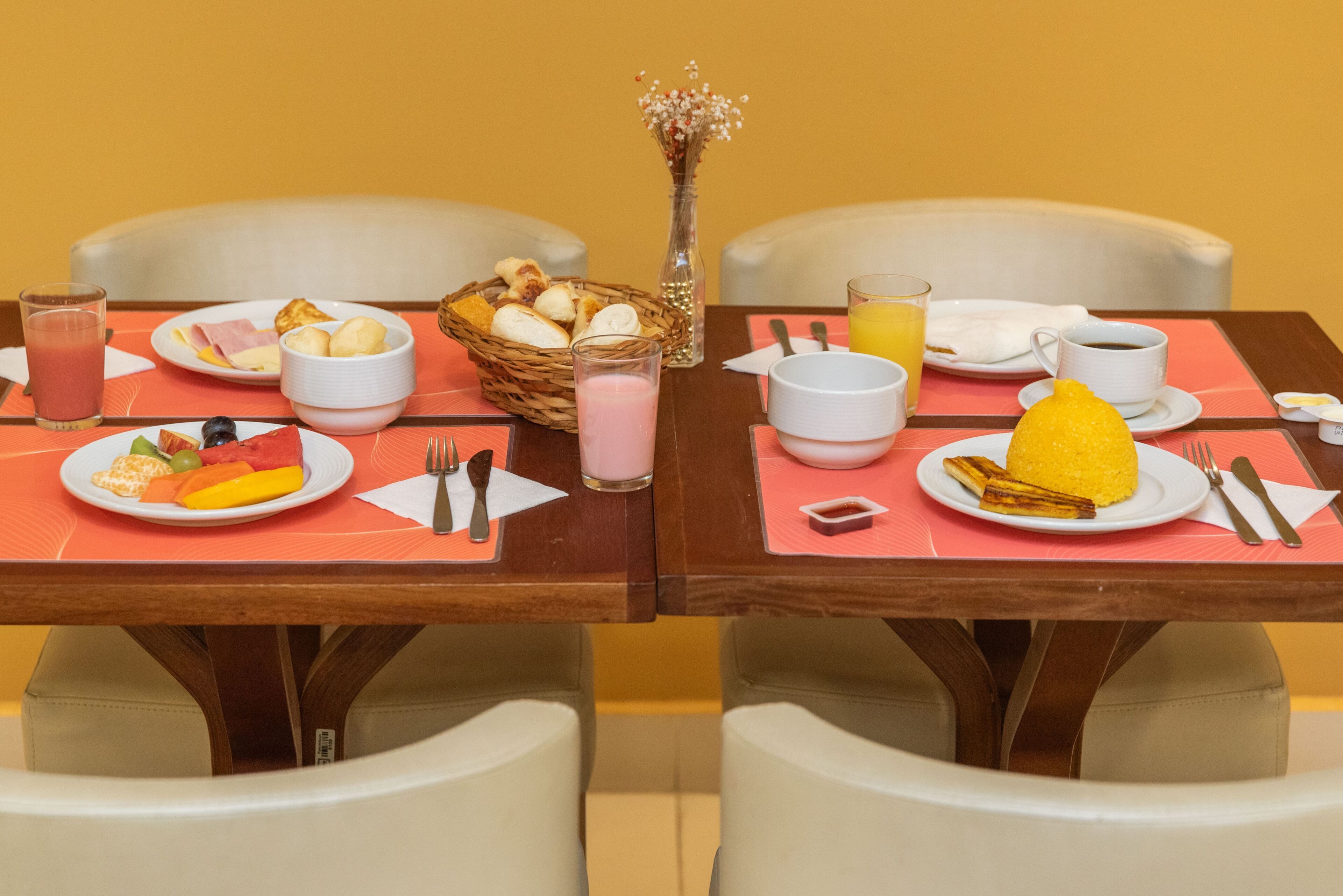 Mesa de café da manhã com pratos de frutas, ovos e torradas, acompanhada por xícaras de café e suco.