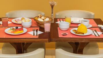 Mesa de café da manhã com pratos de frutas, ovos e torradas, acompanhada por xícaras de café e suco.