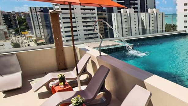 A rooftop pool area with lounge chairs and an umbrella, overlooking a cityscape and a pool. - Verano Pajuçara by Tropicalis (Maceió)