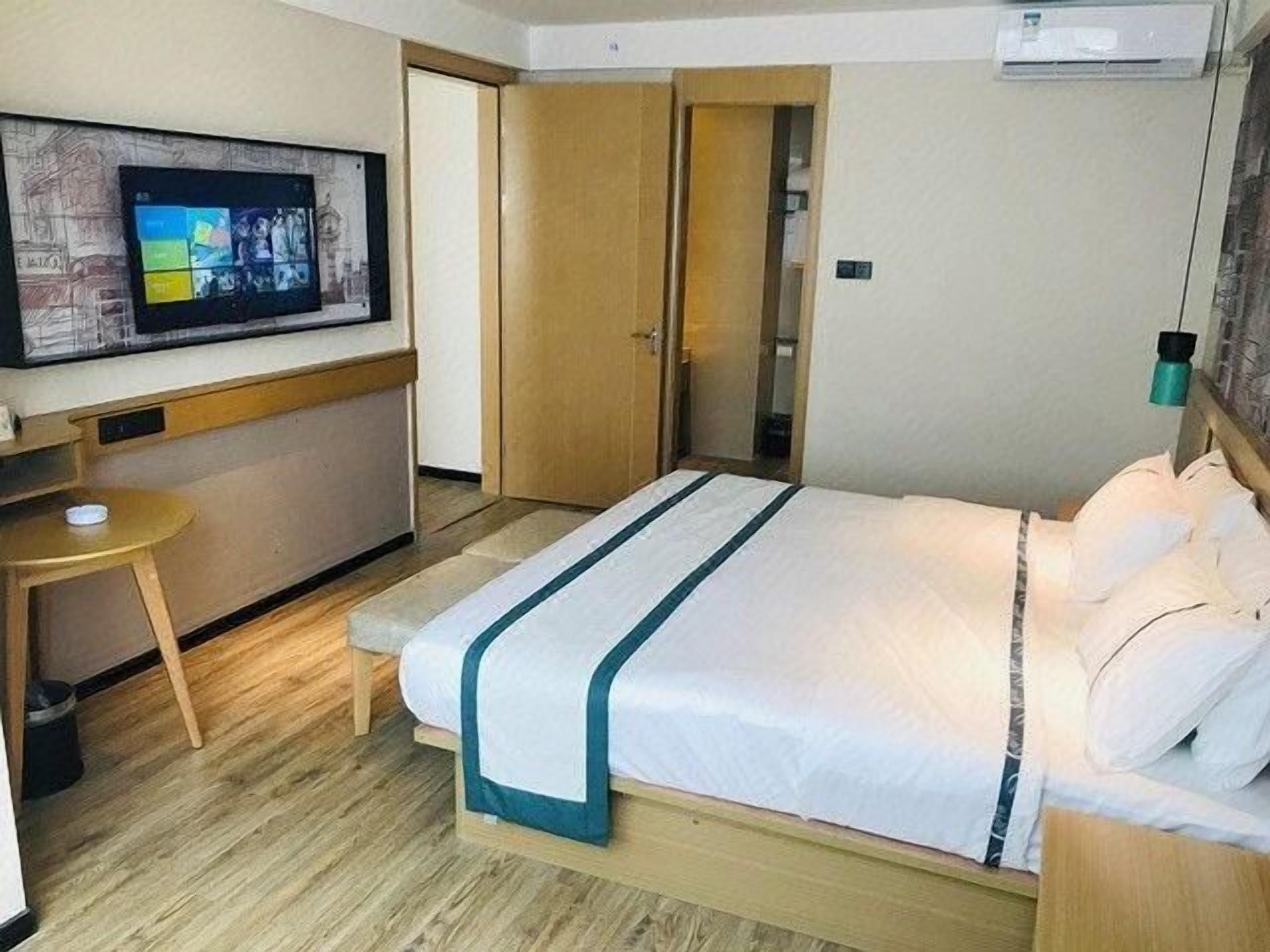 A hotel room with a bed, a TV mounted on the wall, a small table, and a door leading to another room.