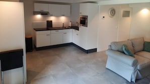 A modern kitchen with white cabinets, a black countertop, and a built-in microwave. There is a beige sofa with green cushions in the living area. - Souterrain "Edelweiss" (Noordwijk)