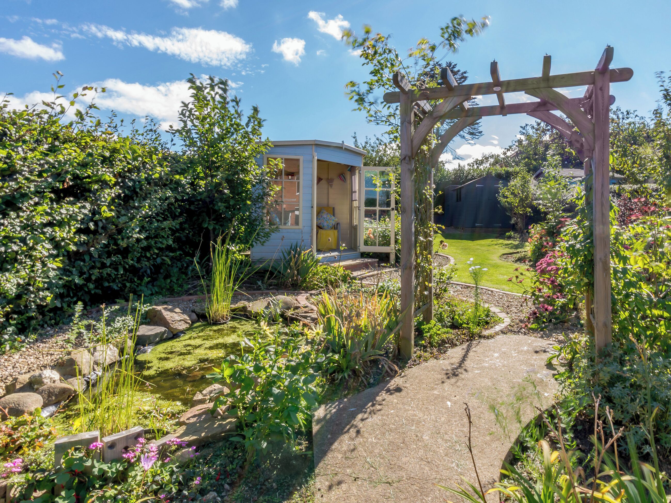 Ein Garten mit einer Holzpergola, einem Teich und einem Haus mit blauer Fassade.