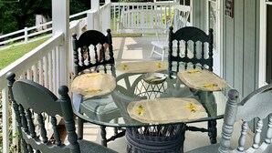 A covered porch with a glass table and chairs, surrounded by white railings and greenery.