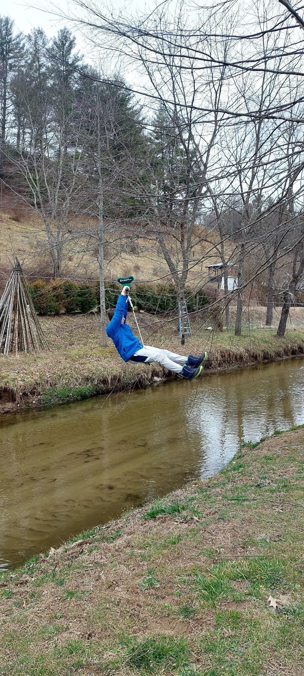 Una persona con chaqueta azul y pantalones blancos sentada en una hamaca junto a un río tranquilo, rodeada de árboles y un área verde.