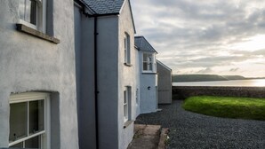 A row of white buildings with grey roofs, a gravel path, and a grassy area leading to a body of water. - Brook Cottage - 1 Bedroom Cottage - Dale (Haverfordwest)