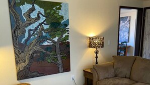En stue med en beige sofa, et sofabord, et træmaleri, en lampe og en mønstret måtte.