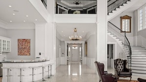 A spacious foyer with a high ceiling, a grand staircase, a central chandelier, and a bar area with stools.