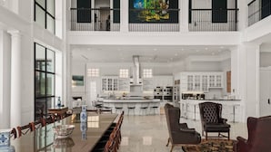 A spacious, high-ceilinged living area with a long dining table, comfortable seating, and a kitchen in the background.