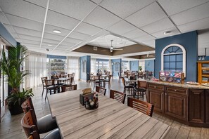 A spacious dining area with multiple tables and chairs, a central island with a beverage cart, and a view of the outdoors through large windows.