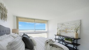 Un dormitorio con cama, ventana con vista a la playa, mesita de noche con lámpara y un cuadro abstracto colgado en la pared.