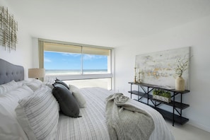 A bedroom with a bed, a window with a beach view, a bedside table with a lamp, and a wall-mounted abstract painting.