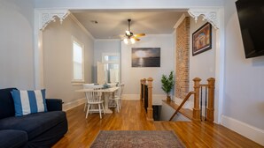 A living room with a dark sofa, a dining area with white chairs, a wooden staircase, and a ceiling fan. - Historic Gem w/ Jacuzzi / Prime Streetcar Spot (New Orleans)