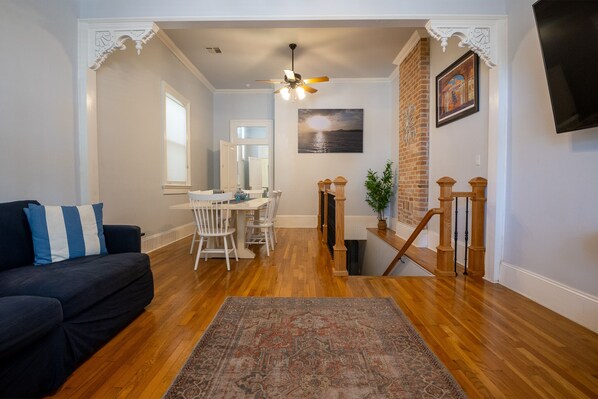 A living room with a dark sofa, a dining area with white chairs, a wooden staircase, and a ceiling fan.