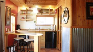 A compact kitchen with wooden cabinets, a sink, and a small refrigerator. There are two stools and a window providing natural light.