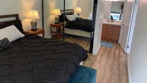 Ein Schlafzimmer mit einem großen Bett, zwei Nachttischen mit Lampen, einem Spiegel, einer Holzbank und Blick auf ein Badezimmer mit Waschbecken und Schränken.