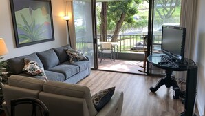 Ein Wohnzimmer mit grauem Sofa, einem beigefarbenen Sessel, einem Fernseher auf einem Schrank und einer Glasschiebetür, die zu einem Außenbereich mit Tisch und Stühlen führt.