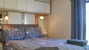 Un dormitorio con cama, almohadas, una mesita de noche, una lámpara y un ventanal con cortinas.