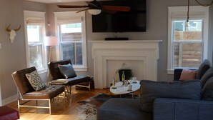 A living room with a fireplace, a television mounted above it, and a ceiling fan. - Cozy Multi-Property Bungalow in Central Austin - Cowboy pool (Austin)